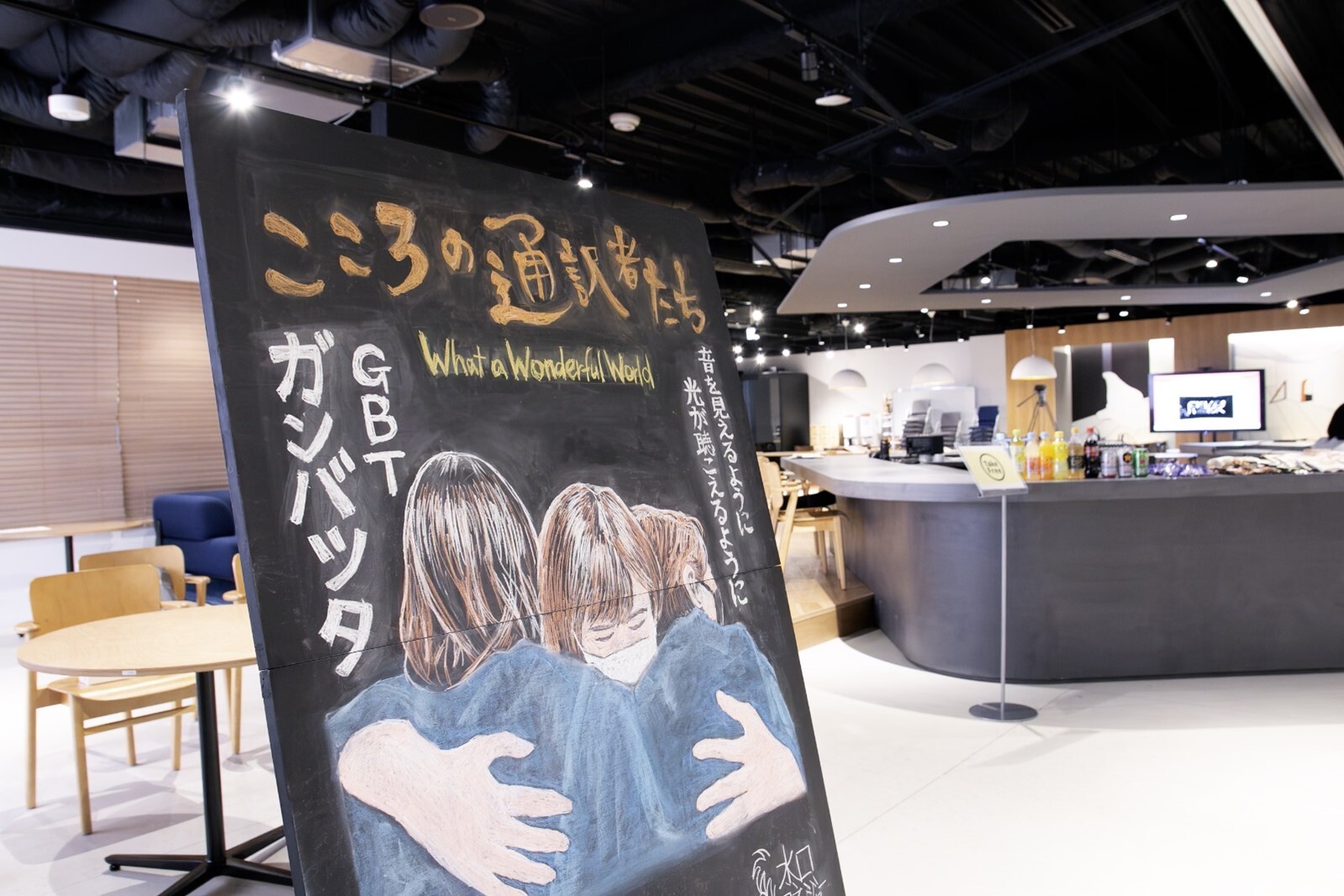 画像 会場に設置された映画『こころの通訳者たち』の黒板アート。タイトルとともに、「音が見えるように、光が聞こえるように」という映画のコピーと、舞台の衣装を着たまま抱きしめ合う手話通訳の3人が、大きく描かれている。イラストの脇には、3人が抱き合いながらつぶやいていた「ガンバッタ」という言葉と、舞台『凛然グッドバイ』で印象的なセリフ「What a Wonderful World」という言葉が。 画像終わり