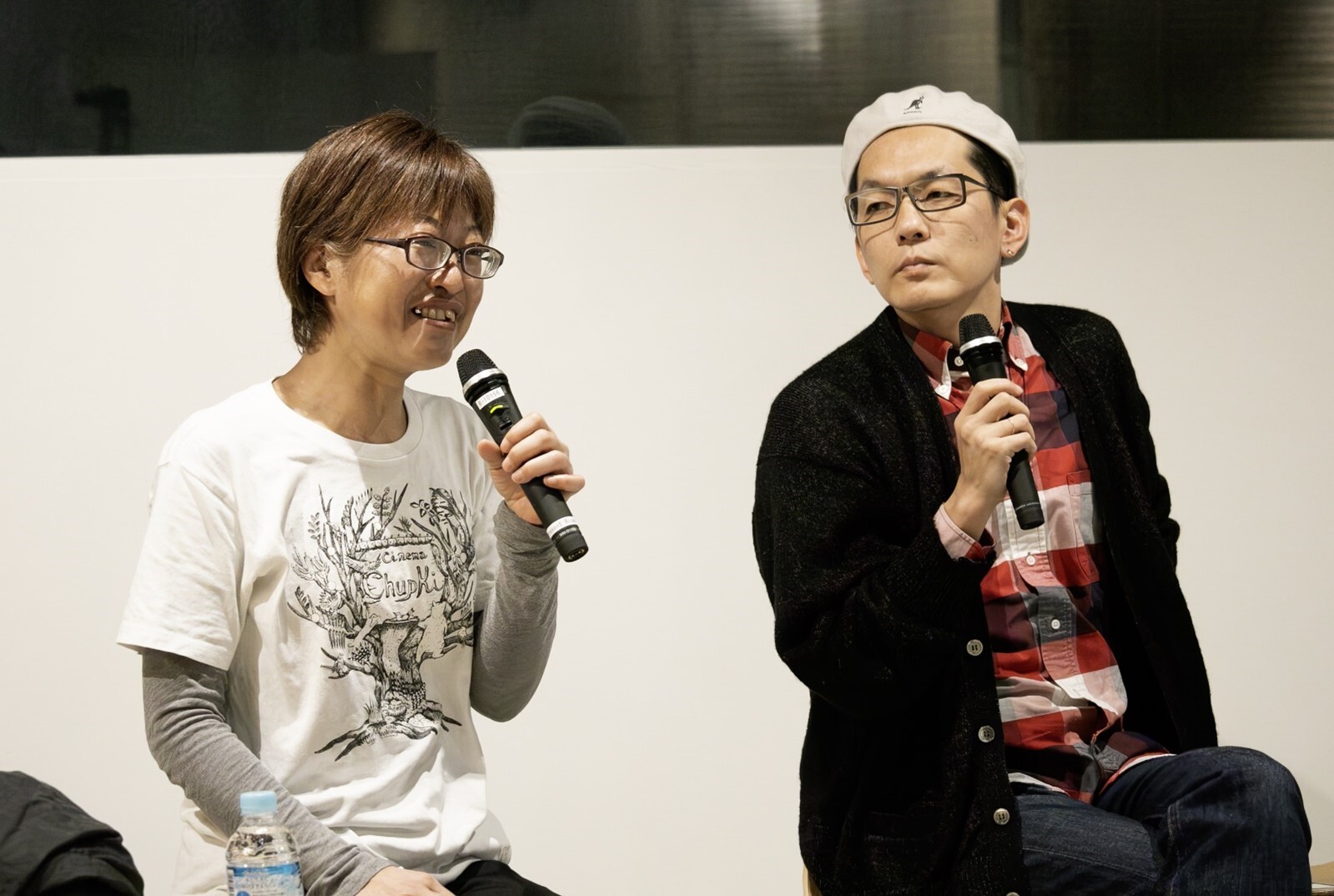 画像 シネマチュプキタバタのロゴが入った白いTシャツで、微笑みながらマイクを持つ平塚さんと、赤と黒と白の3色を基調としたチェックシャツに黒いカーディガン、白のハンチングをかぶり平塚さんのお話に真剣に耳を傾ける石井さん。 画像終わり