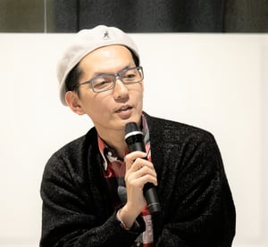 画像 赤と黒と白の3色を基調としたチェックシャツに黒いカーディガン、白のハンチングをかぶり、マイクを持つ石井さん。 画像終わり