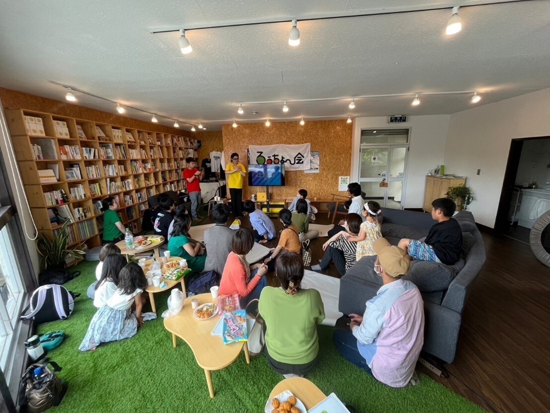 画像：ろうちょ～会さんの普段の活動風景。芝生のような絨毯のある、温かみのある部屋で、年齢や性別を問わずたくさんの方が、参加している。画像終わり