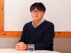 画像:深い紺色のセーターの中にシャツを着てほほ笑む、五十嵐さんのインタビュー中の写真。画像終わり