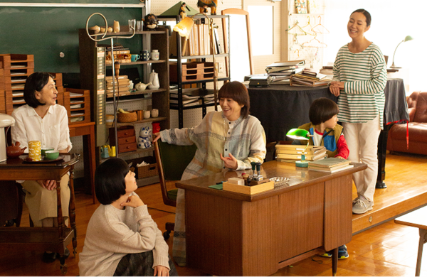 画像：とりつくしまの劇中写真。メインキャストの小泉今日子さんを中心とした登場人物が、学校の教室にも見える、温かみのある部屋に集まり笑い合っている。画像終わり