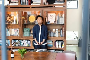 画像：シモキタエキマエシネマK2の大高さんの写真。大きな木製の本棚の前に佇む。心地良い光が差し込み、木のぬくもりのあふれるお部屋で。画像終わり