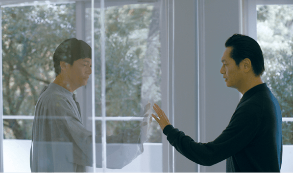 画像：徒花の劇中写真。メインキャストを演じる井浦新さんが、同じく井浦さんが演じているものの、服装も髪型も違う別の存在とガラス越しに手を合わせている。画像終わり