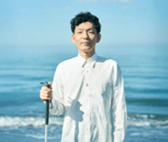 画像：ブラインド・コミュニケーターの石井さんの写真。美しい青い空と海をバッグに、白いシャツを着て白杖を持ち柔らかな表情で正面を見つめる。画像終わり