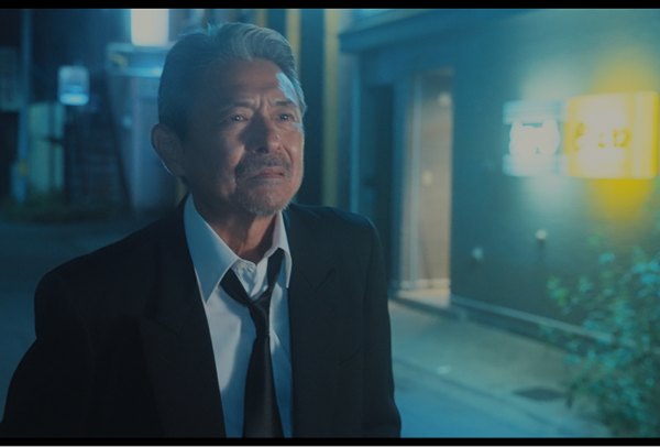 画像：美晴に傘をの劇中写真。夜の街を背景に、升毅さん演じる男性が喪服にも見えるスーツ姿で、何かをこらえるような表情をしている。画像終わり