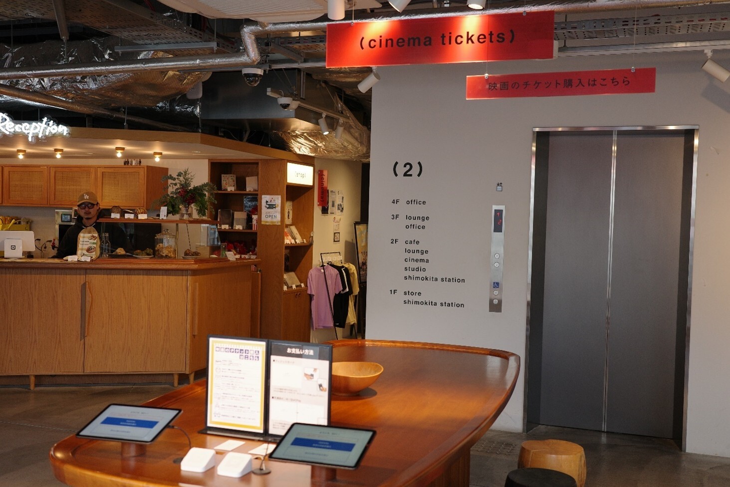 画像：シモキタエキマエシネマK2の入り口の写真。チケット発券機があるが、無人で、タブレットが置いてある。全体的に木目の落ち着く空間。画像終わり